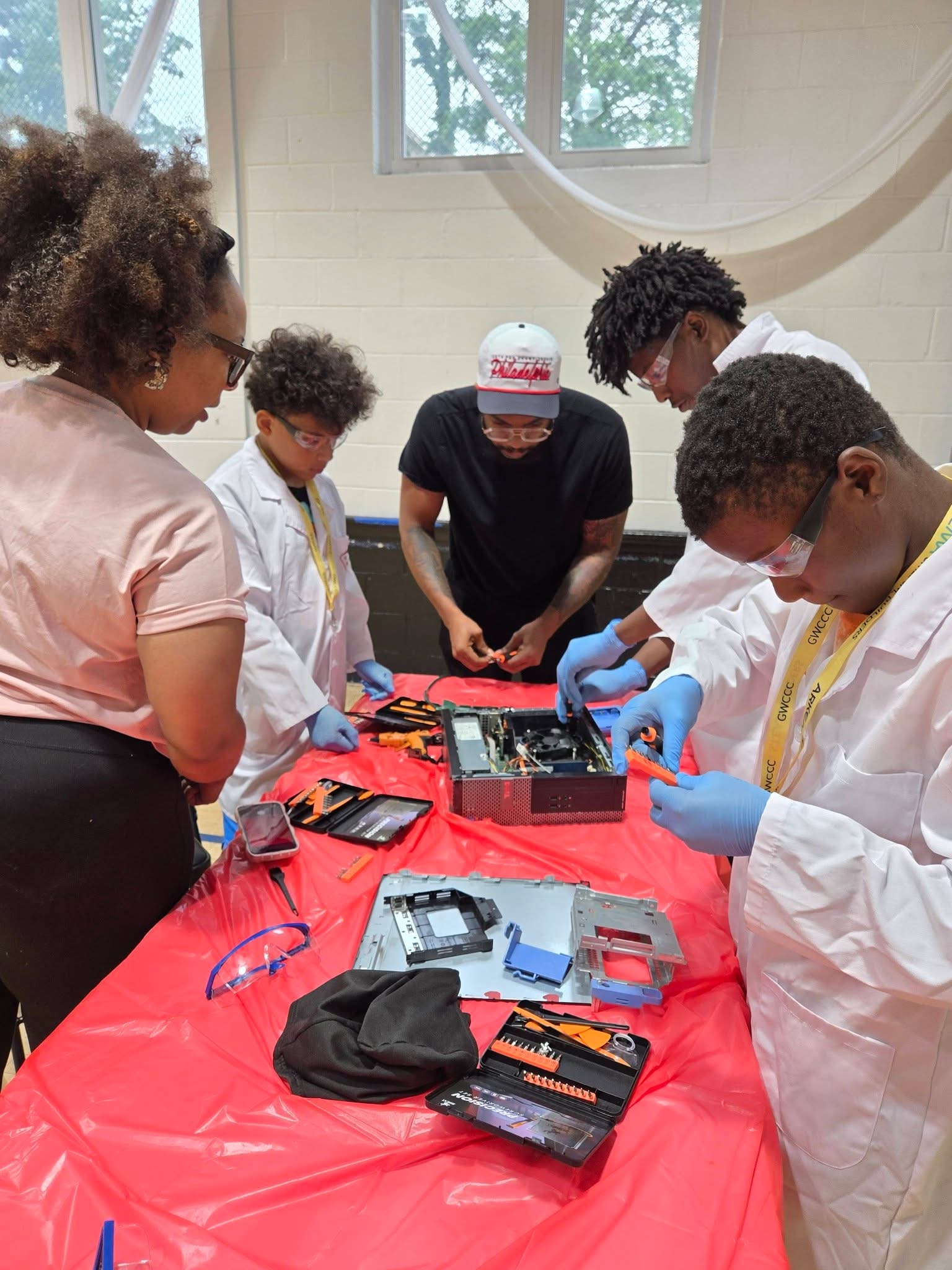 Students in lab coats and gloves work together on a disassembly at a shared table.