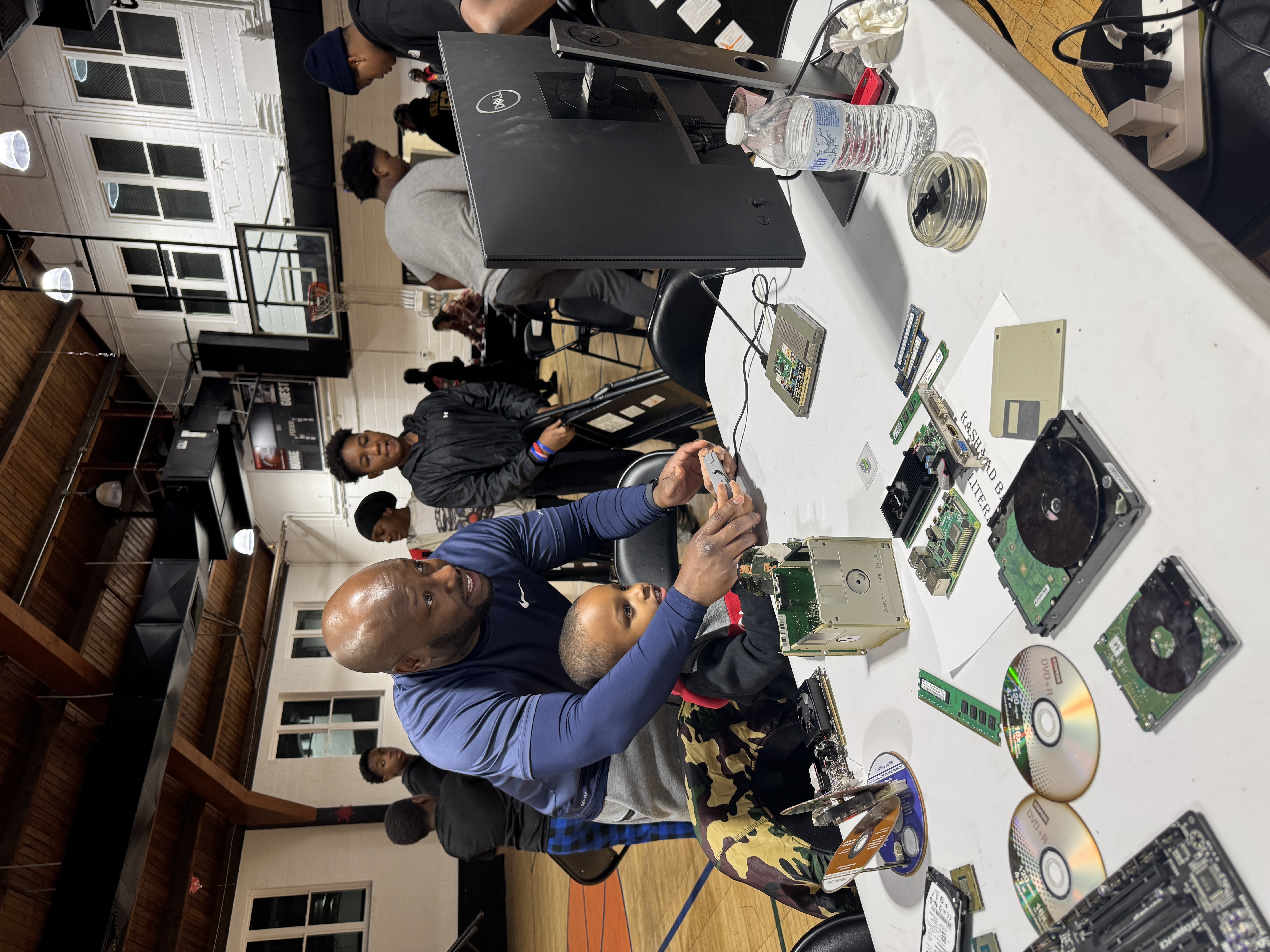 A technician works through a stack of hard drives and components at a repair bench with students looking on.