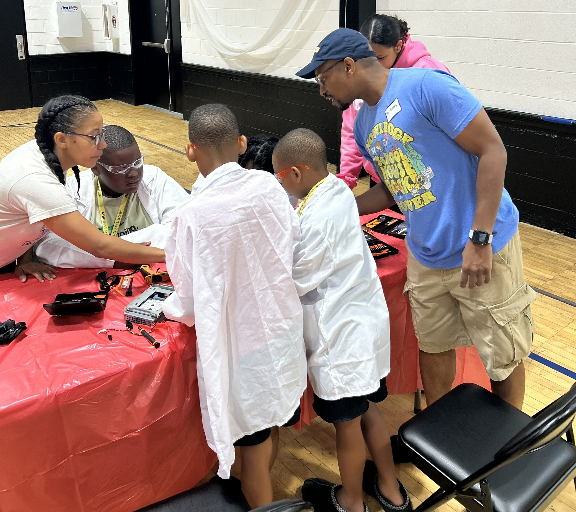 A mentor leans over a workshop table guiding four students in lab coats through a computer disassembly.