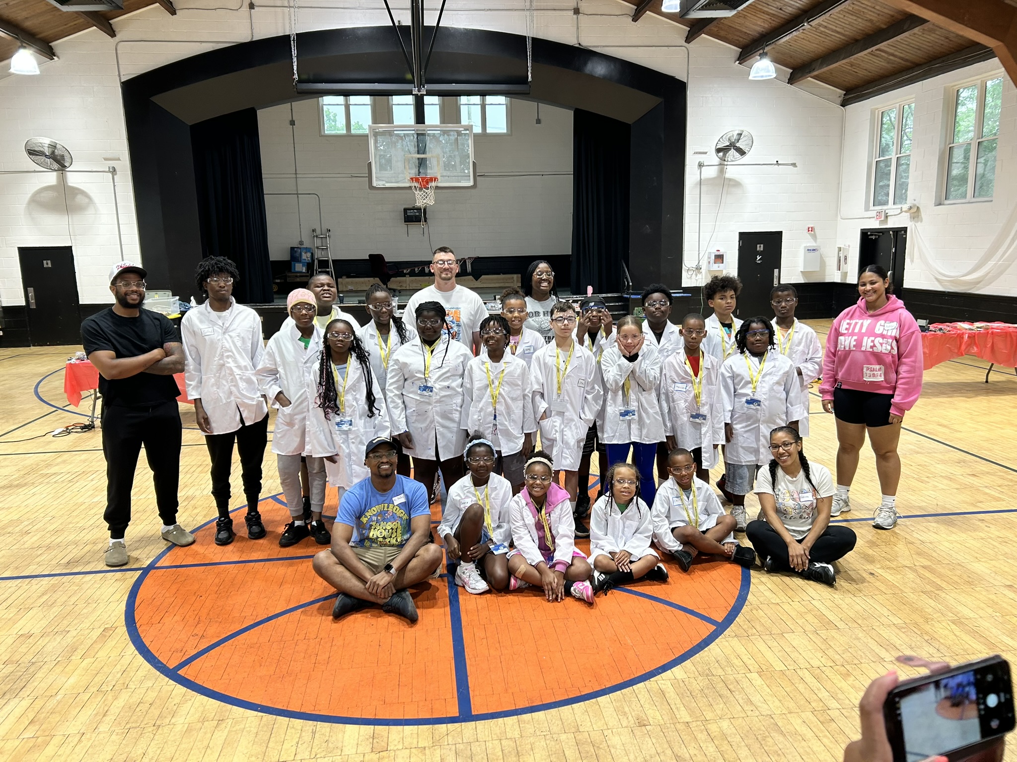 A large group photo of students in white lab coats with their mentors in a community centre gym.