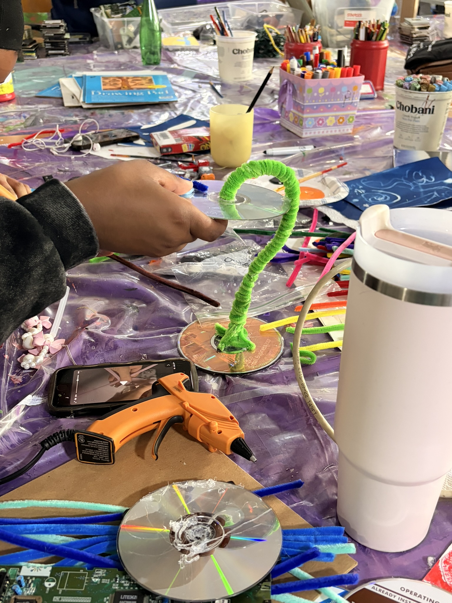Hands assembling a sculpture from a CD, a circuit board and green pipe cleaner at a craft table.