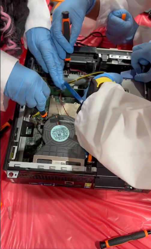 A young boy in goggles uses a hand tool to open a hard drive case at a workshop table.